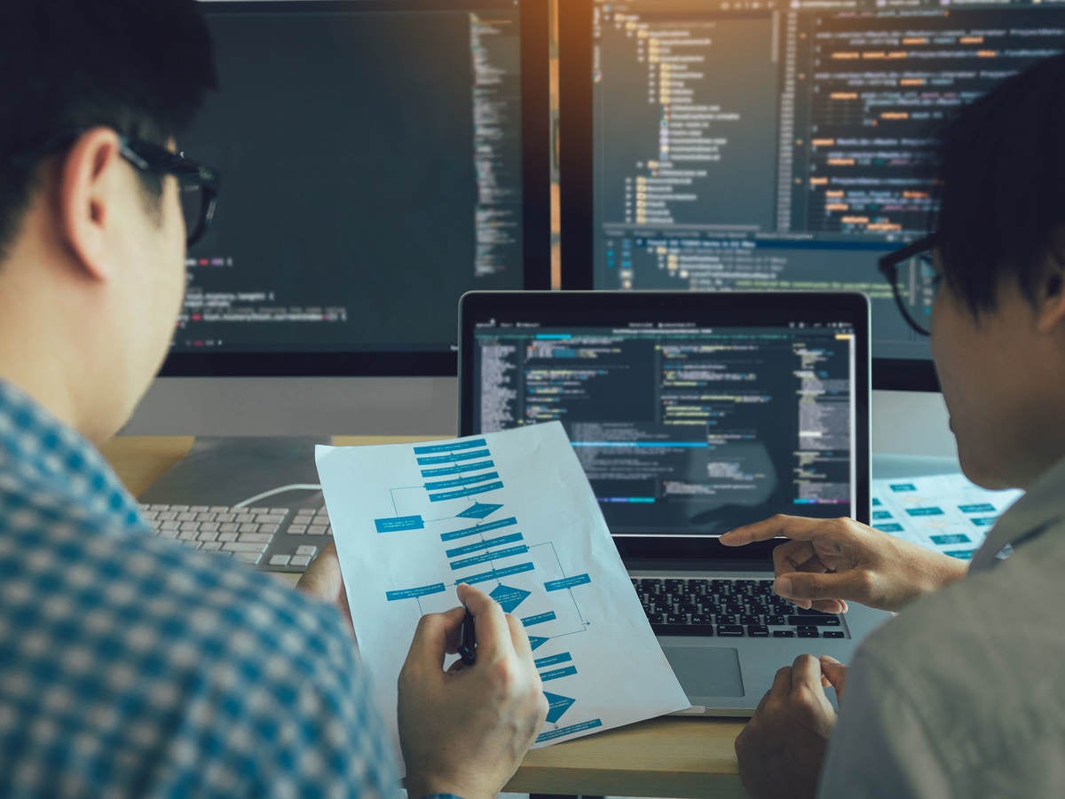 Two engineers examining a process flow on paper with coding on computer screens in the background.