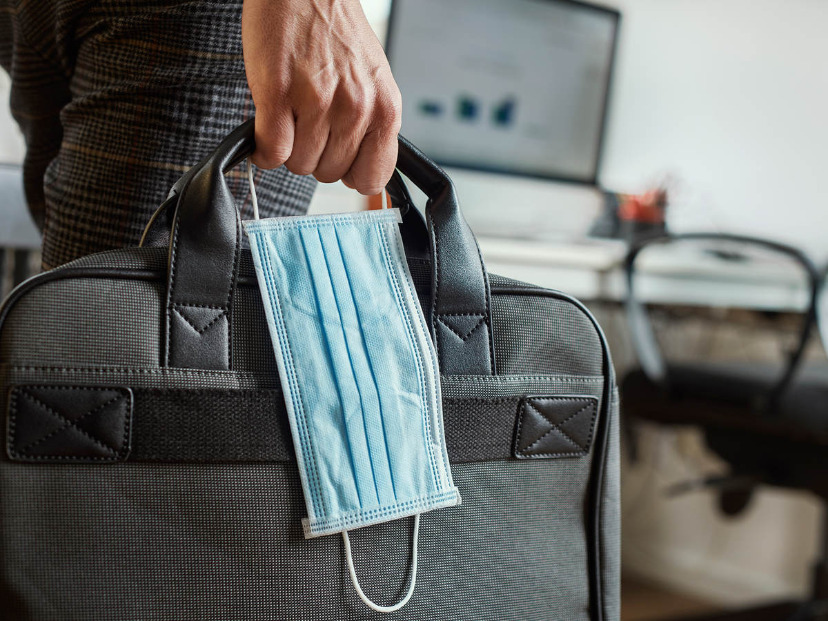 Man in an office holding a briefcase and surgical mask in his hand