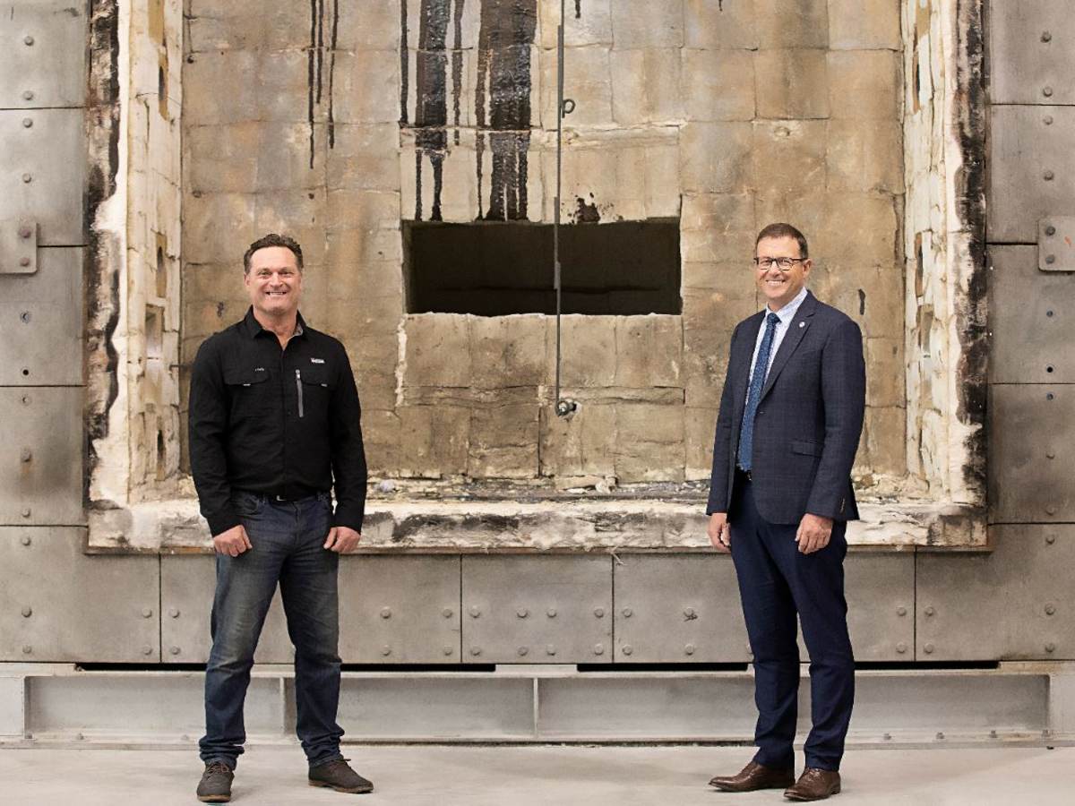 Two white males, one in a black sweater and jeans, and one in a suit and tie, stand smiling in front of a fire test furnace