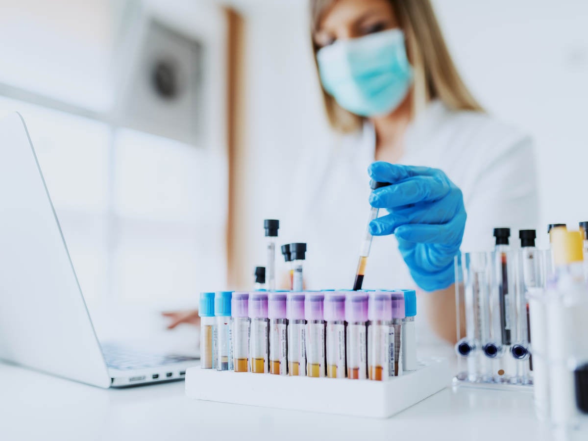 Woman wearing face mask in bright white lab observing vials while logging information on laptop