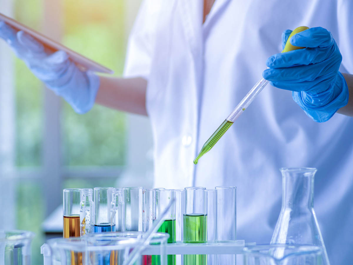 Laboratory worker with blue gloves holds a pipette with green liquid over a several test tubes