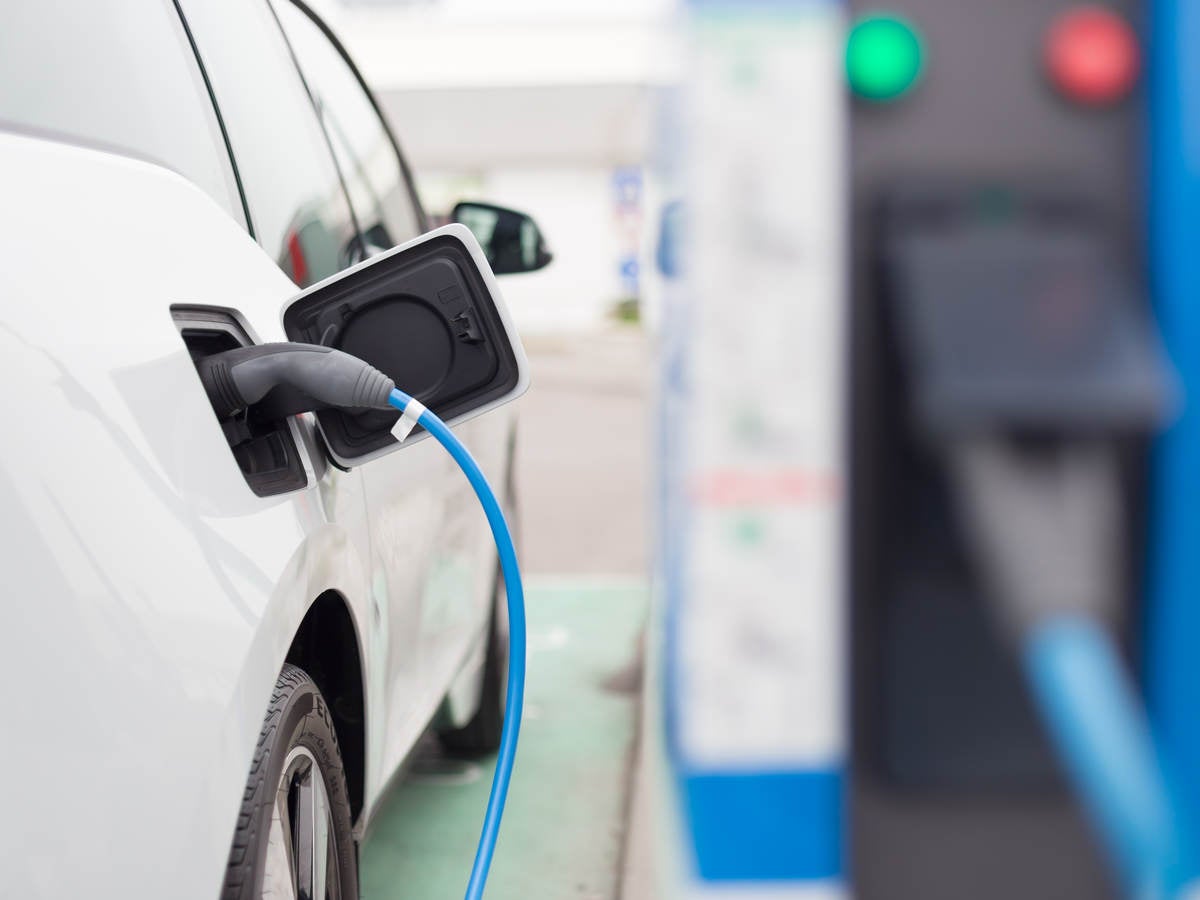 White electric car with charging cord attached; charging station in right background