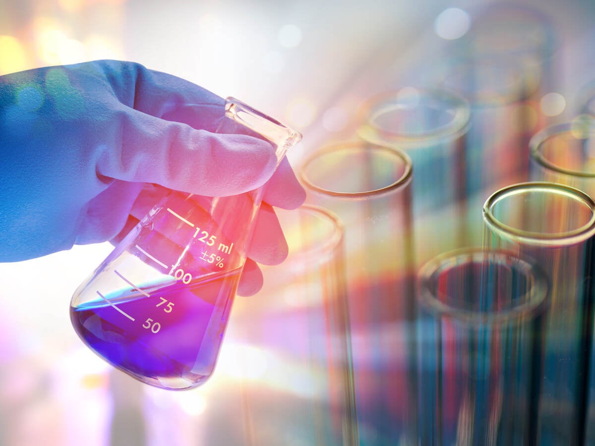 Rubber-gloved hand holding beaker containing blue liquid on a colorful background with test tubes