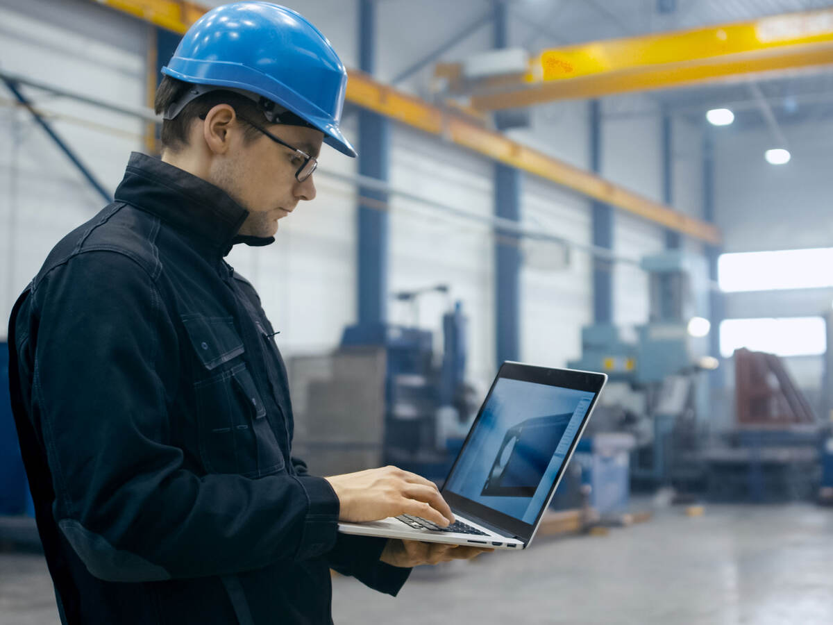 Factory worker in a hard hat is using a laptop computer with engineering software