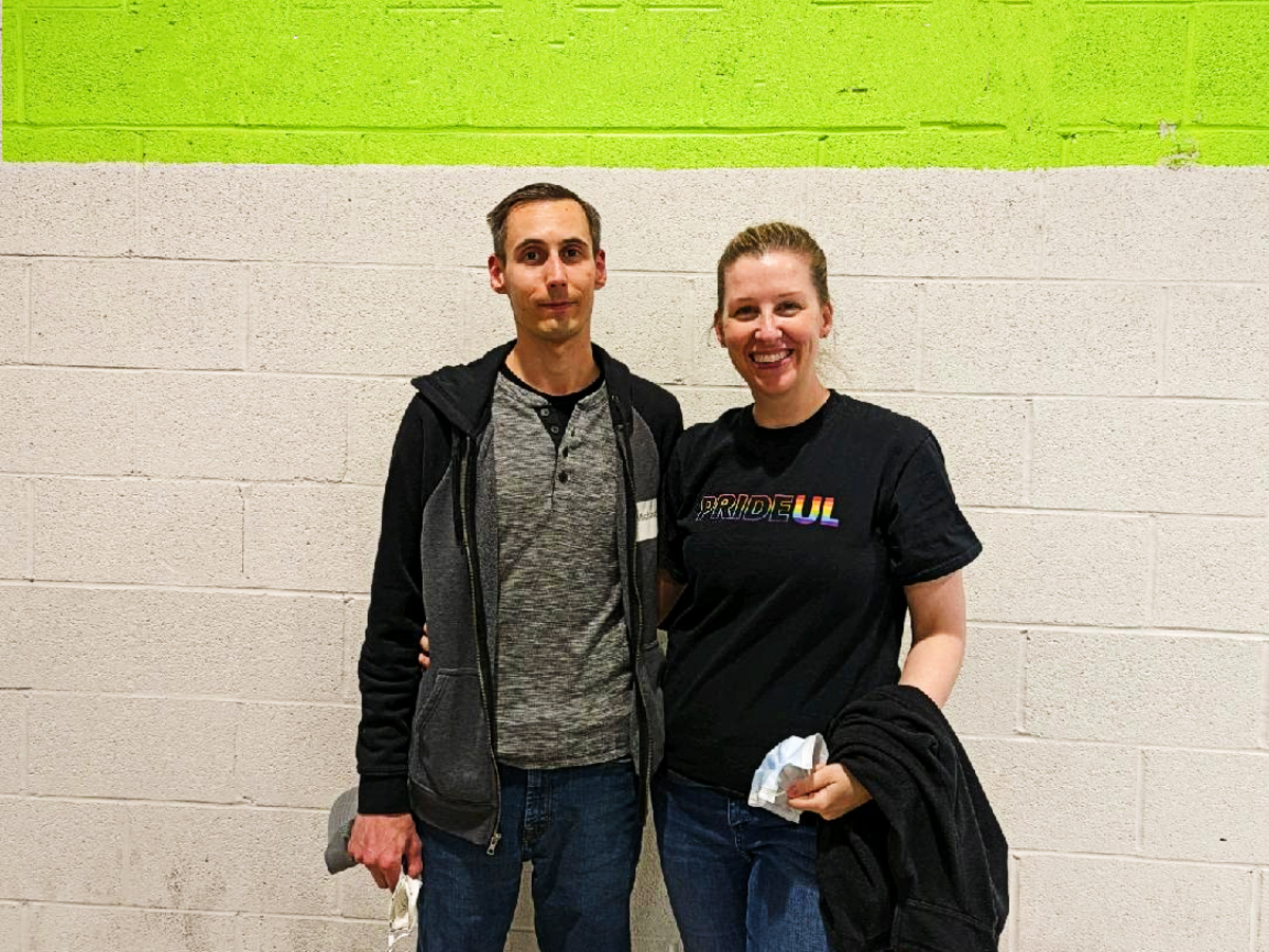 Man and woman from UL PRIDE smile while standing in front of white wall at volunteer event. 