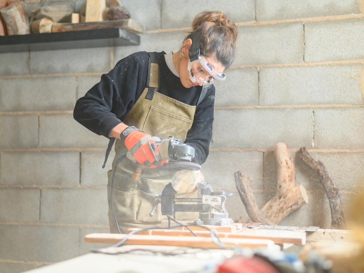 A carpenter, using proper safety equipment, rounds a piece of wood with an angle grinder.