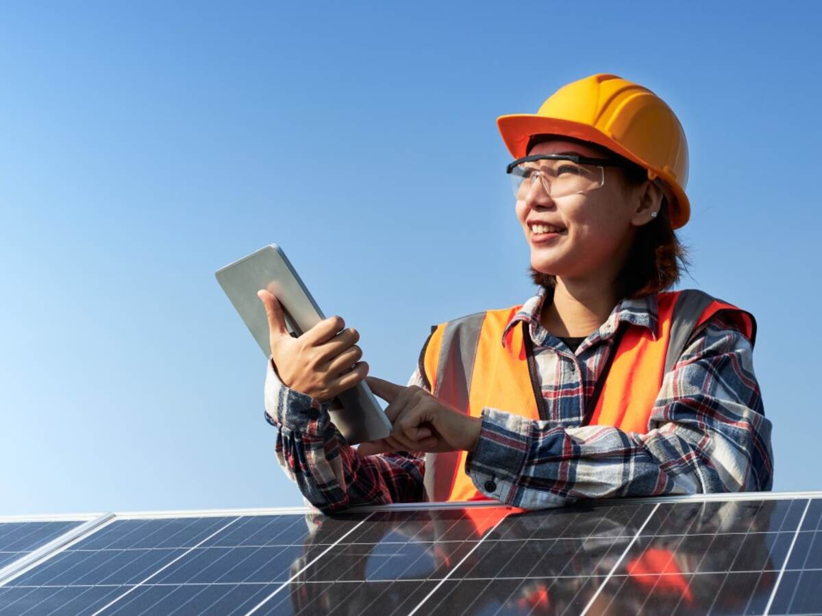 Female engineer in orange safety gear tests solar panel with tablet. 