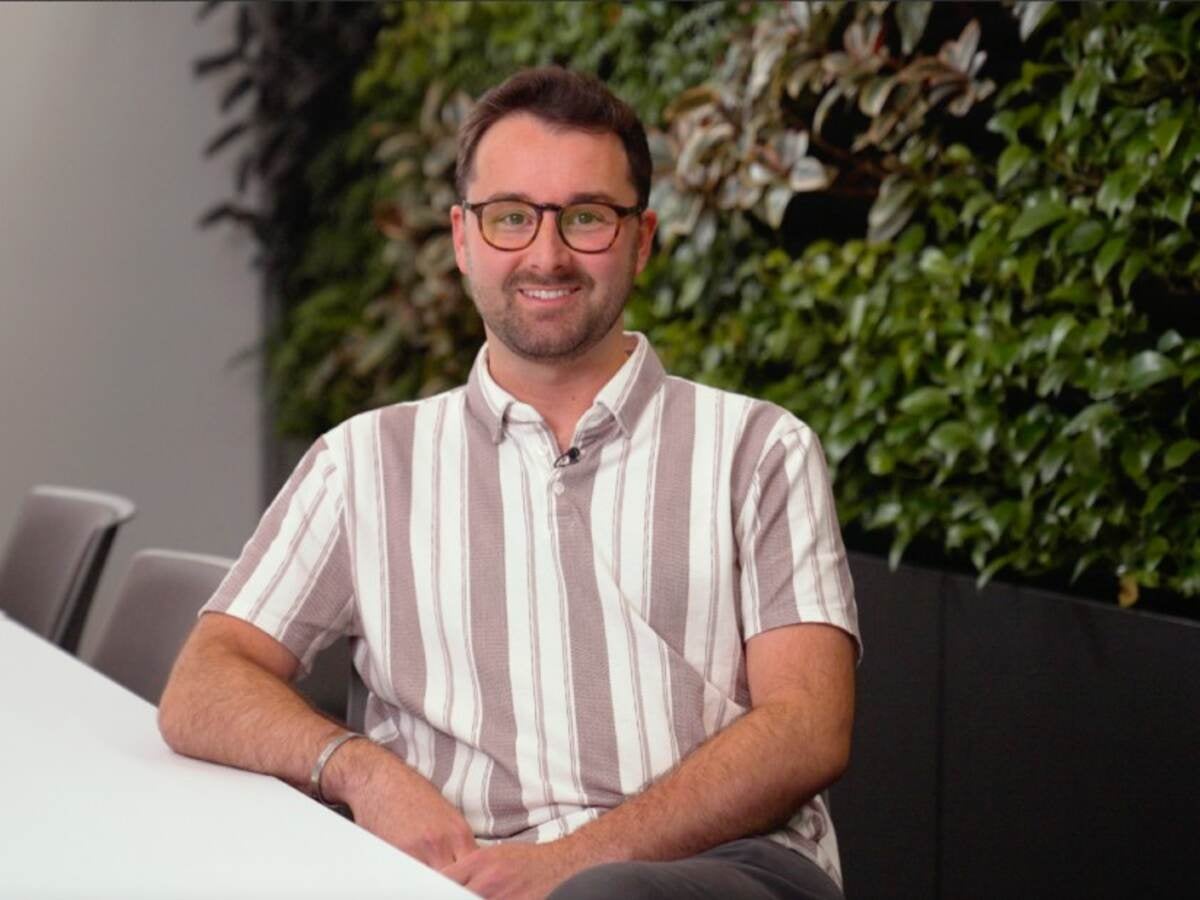 Jacob Tenuta wearing striped shirt sits in front of wall of greenery. 