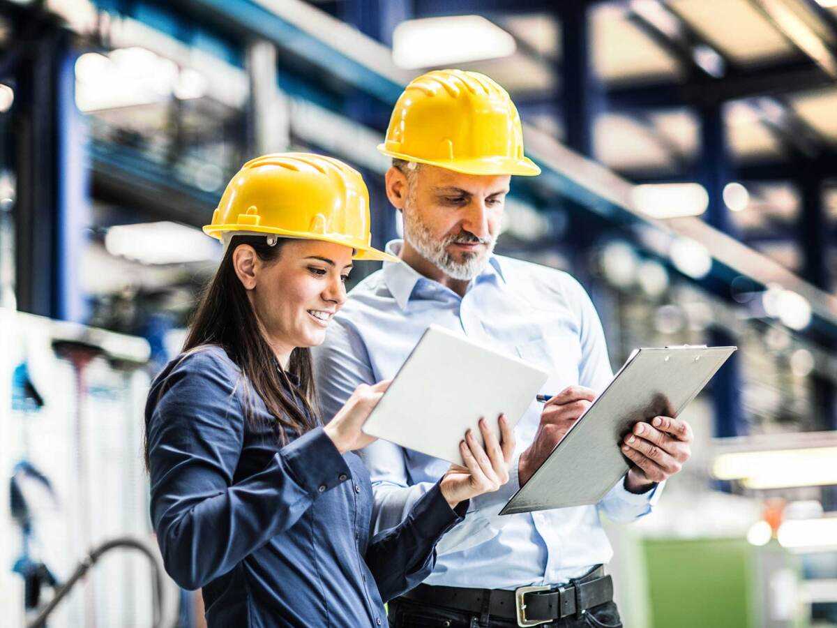 Two workers in yellow hardhats comparing notes on their tablet and clipboard 