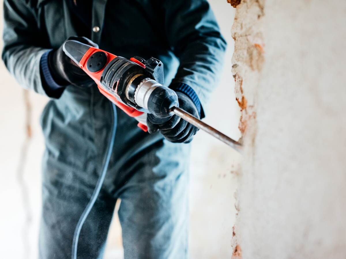 Close up of man's hands holding drill and demolishing wall at house.