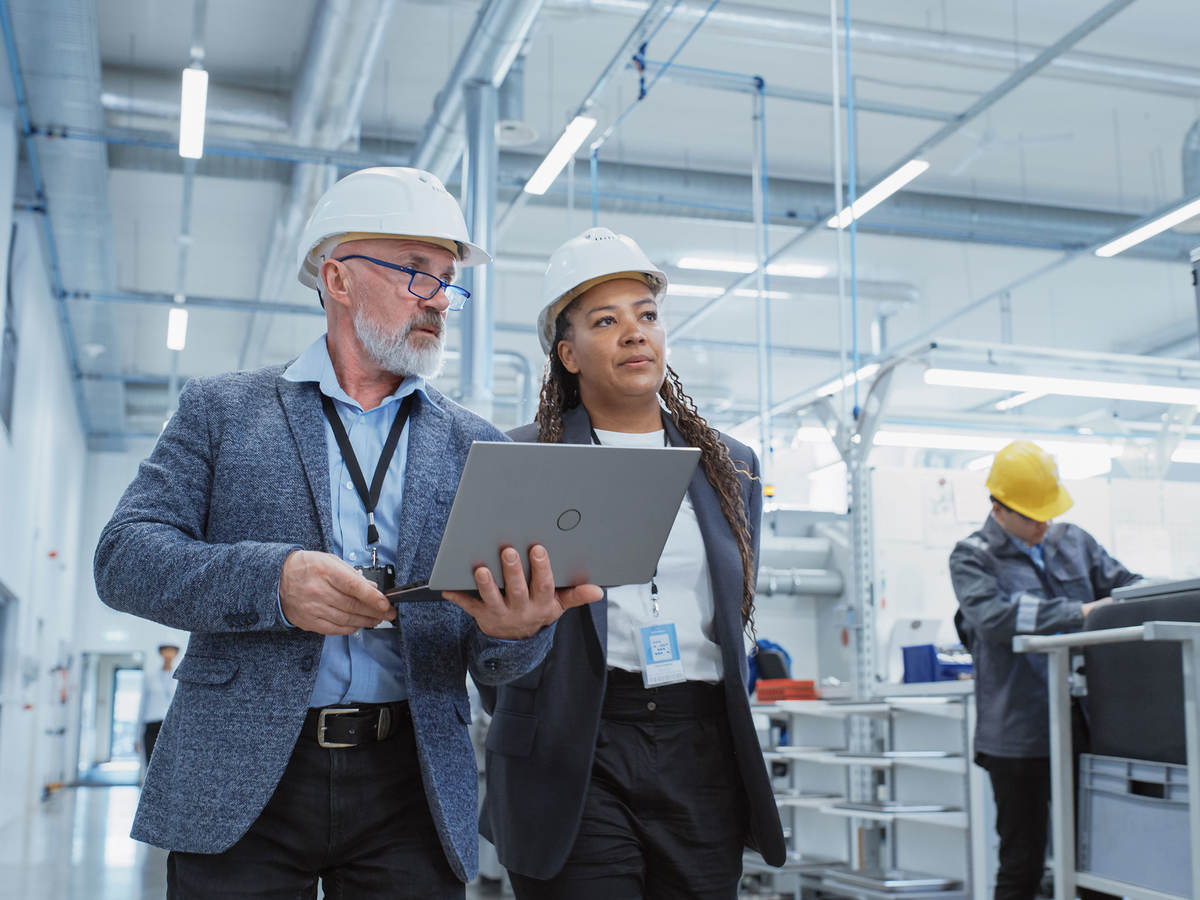 Two managers touring a factory floor while reviewing a laptop