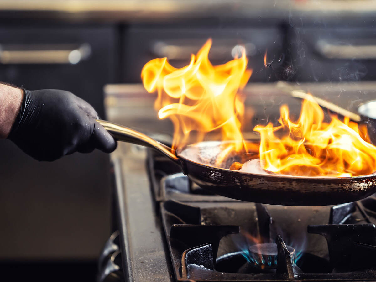 A chef wearing a protective glove while holding a frying pan that has flames coming off of it