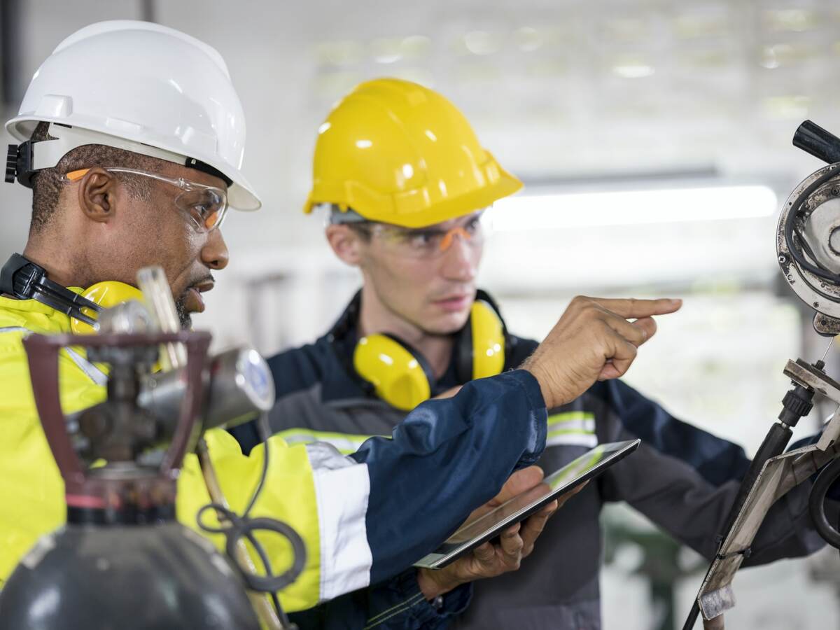 Factory line workers in safety gear examine and discuss a machine part.