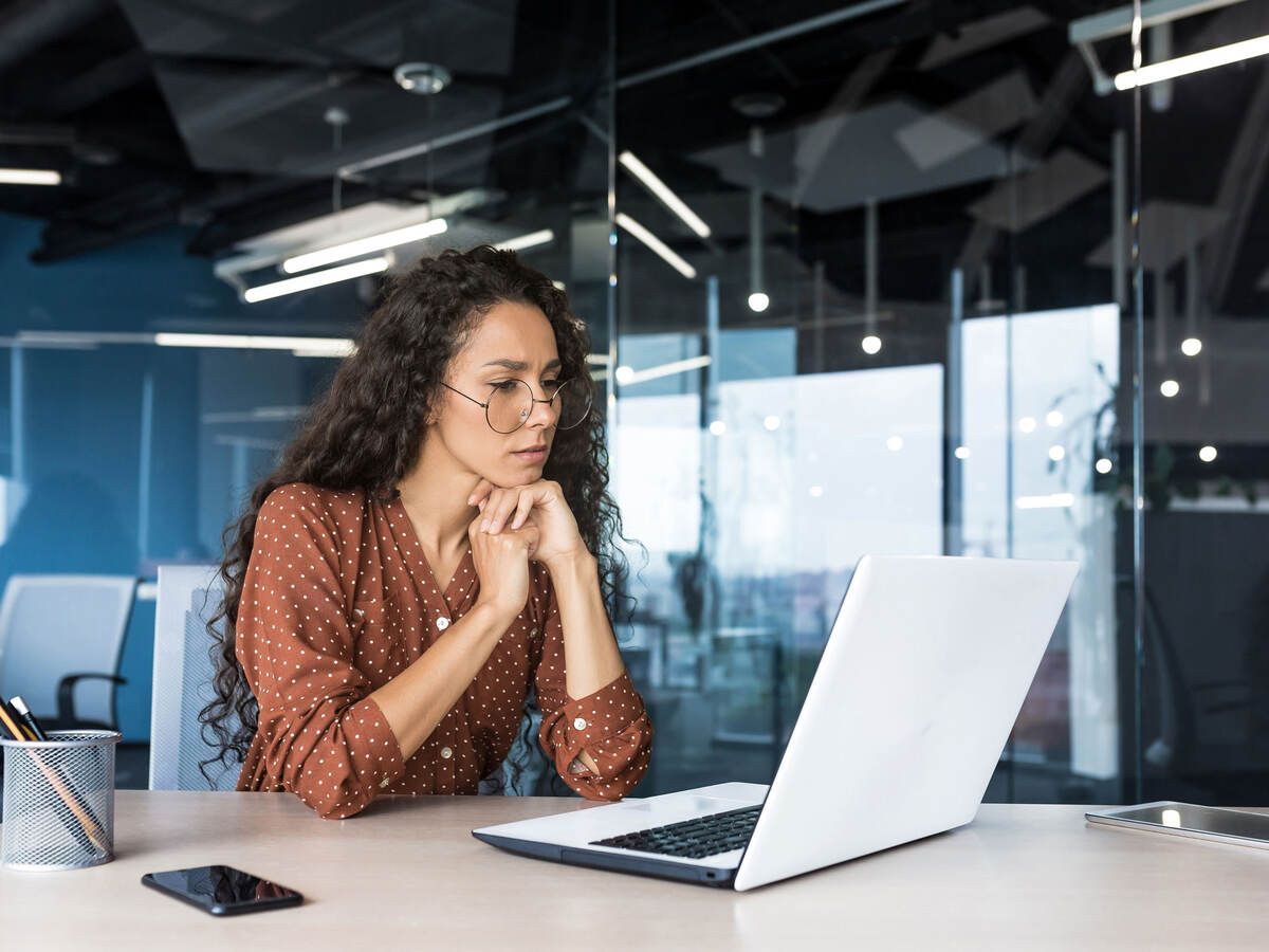 Person in a glass office, looking thoughtfully at a laptop