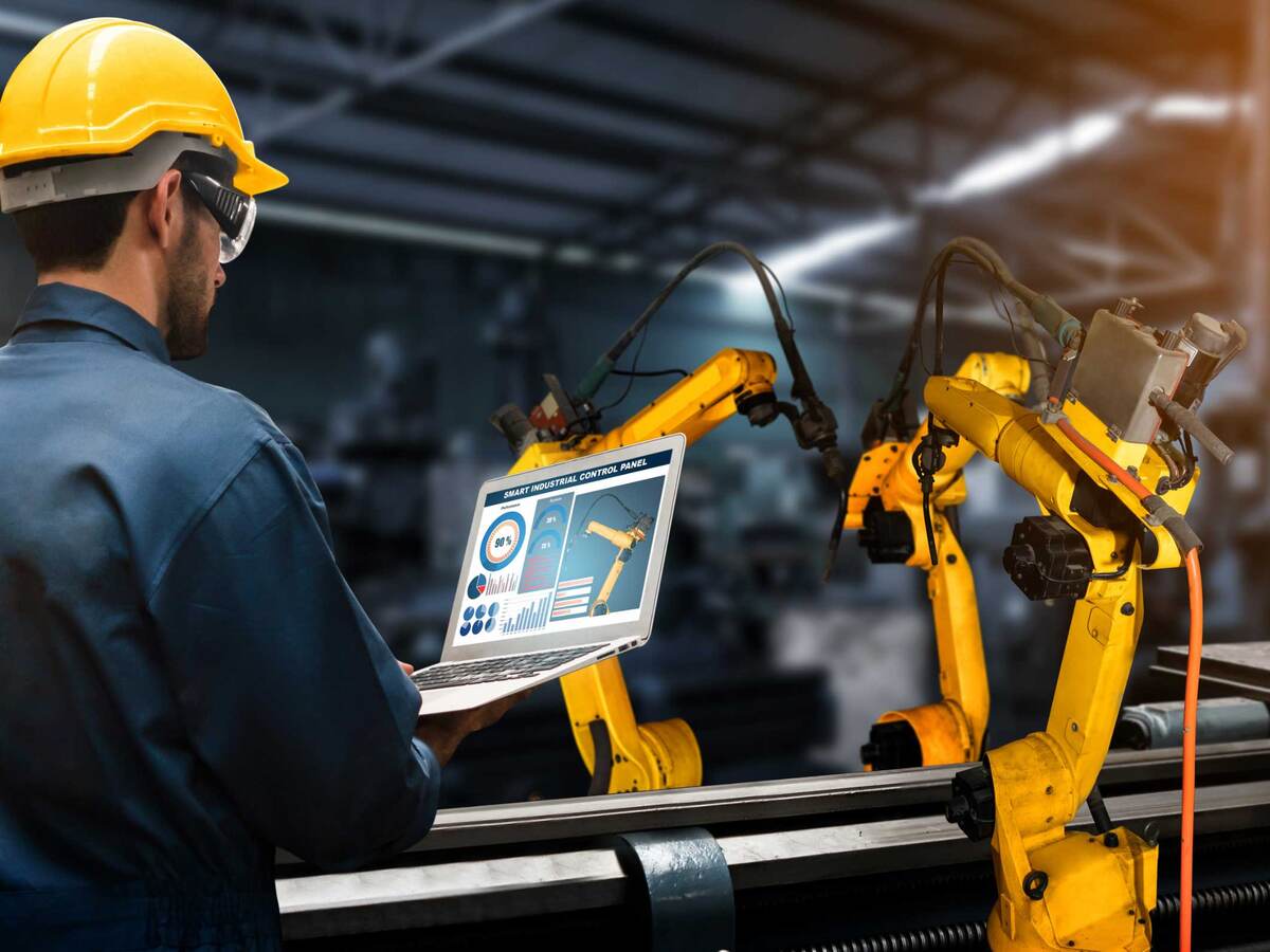 Engineer looking at a laptop in front of and assembly line controlled by robotic arms