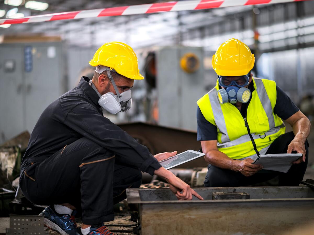 Two officers wearing gas masks, holding tablet and book, inspect the chemical spill