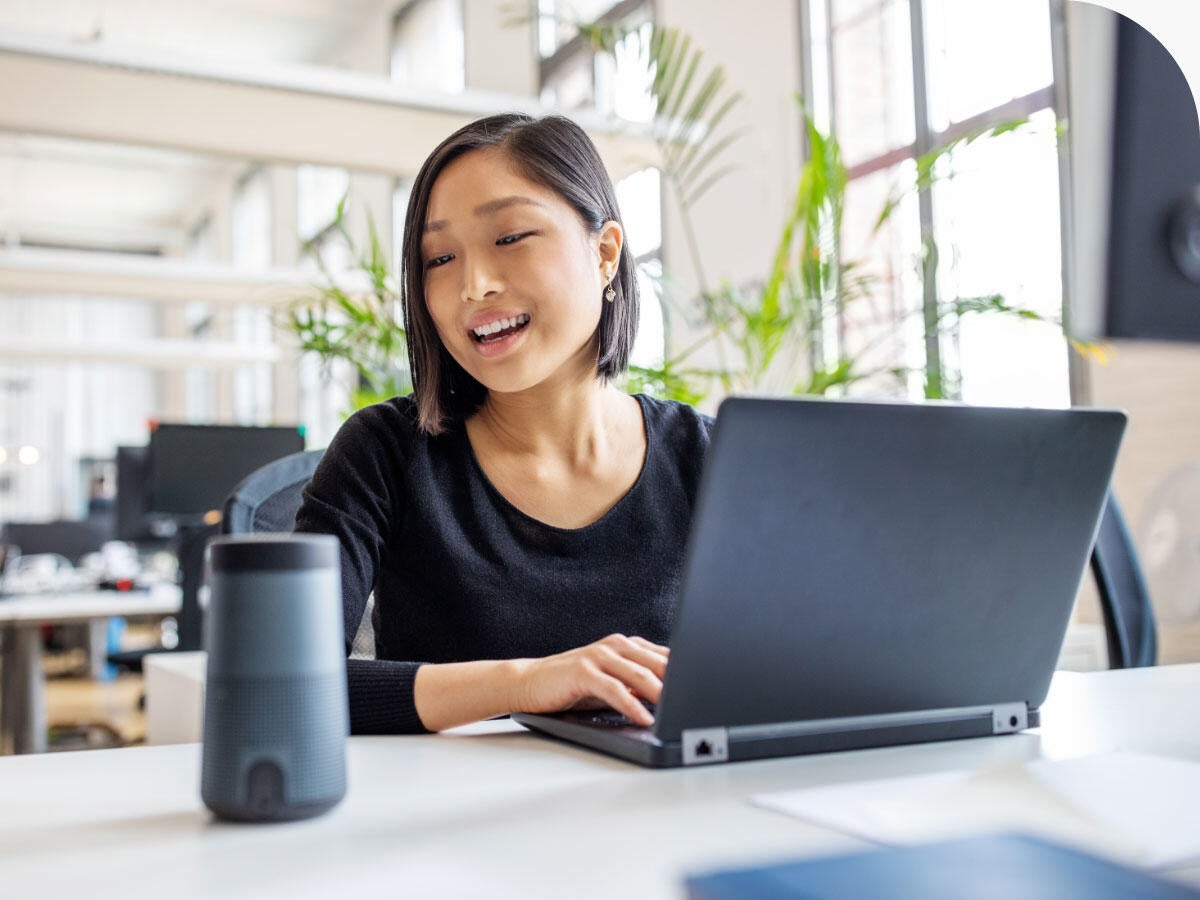 Business professional working on laptop and talking to a virtual assistant via smart speaker