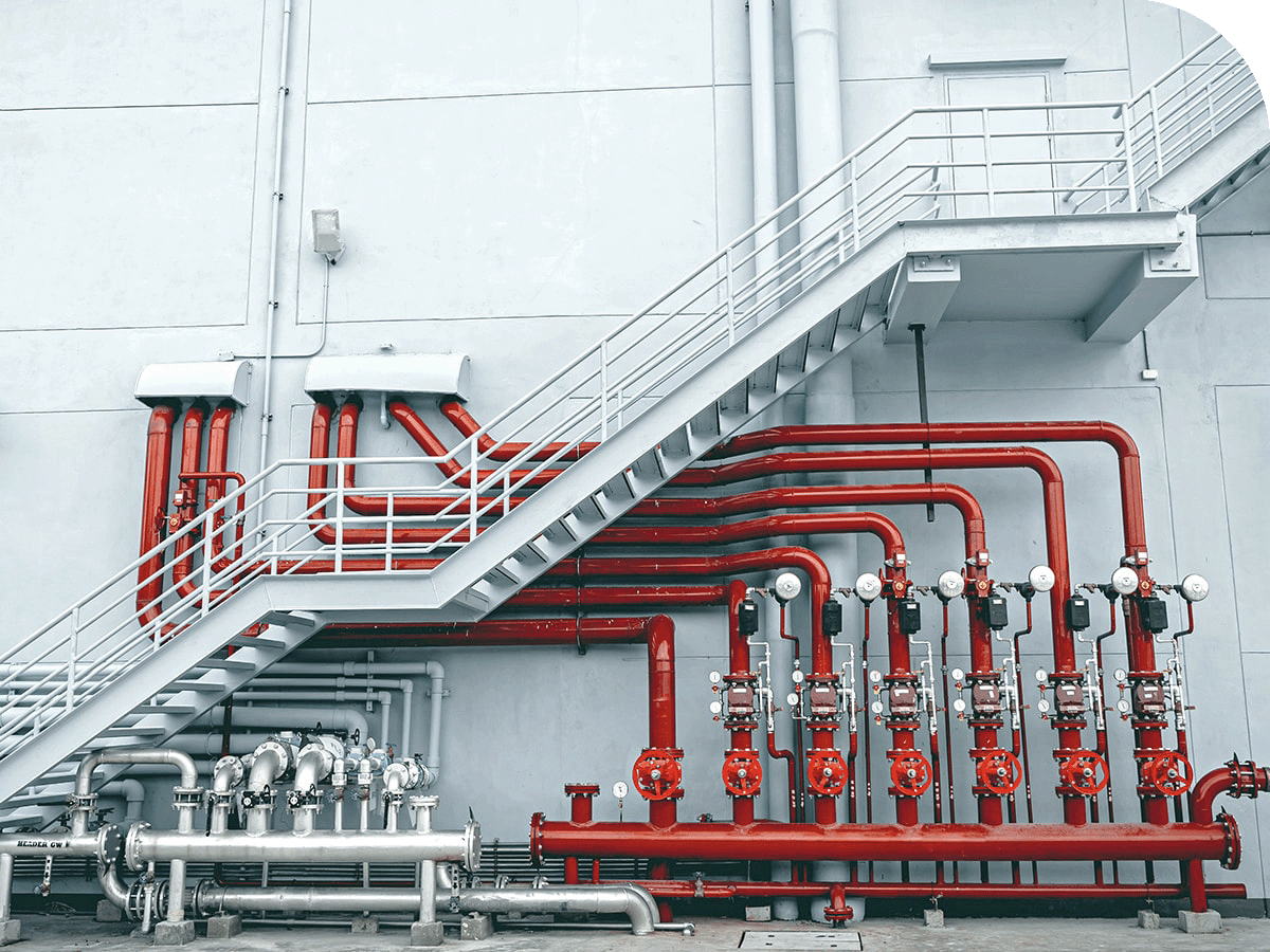 Red industrial pipes and valves mounted on a gray wall beneath a metal staircase.