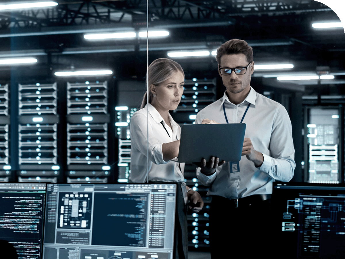 Two people standing in a server room, reviewing information on a laptop, with multiple monitors displaying code and data in the foreground and server racks in the background.