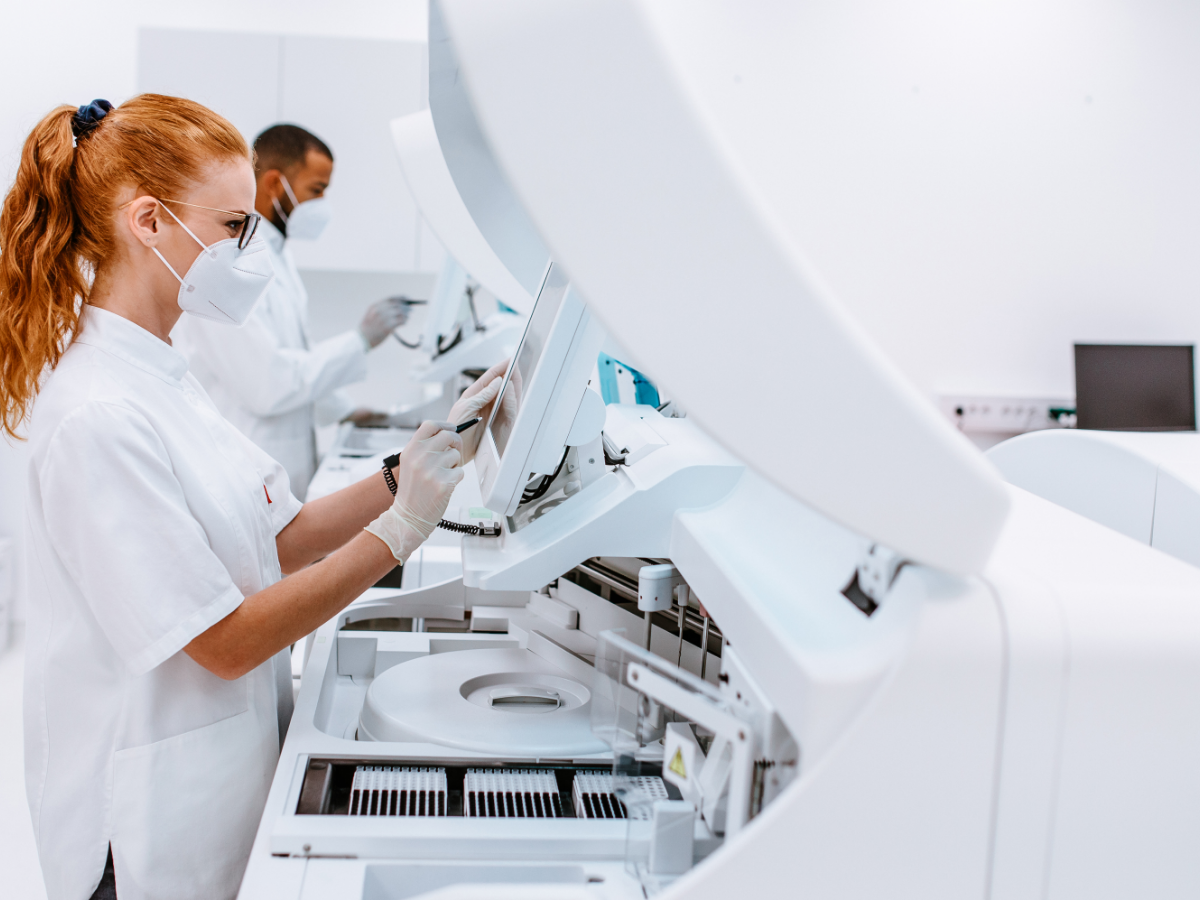 Two laboratory professionals wearing white lab coats, face masks, and gloves work in a modern, sterile lab. The woman in the foreground interacts with a large automated diagnostic machine via a touchscreen, while the man in the background operates another piece of equipment. The setting highlights advanced medical technology and strict hygiene protocols.