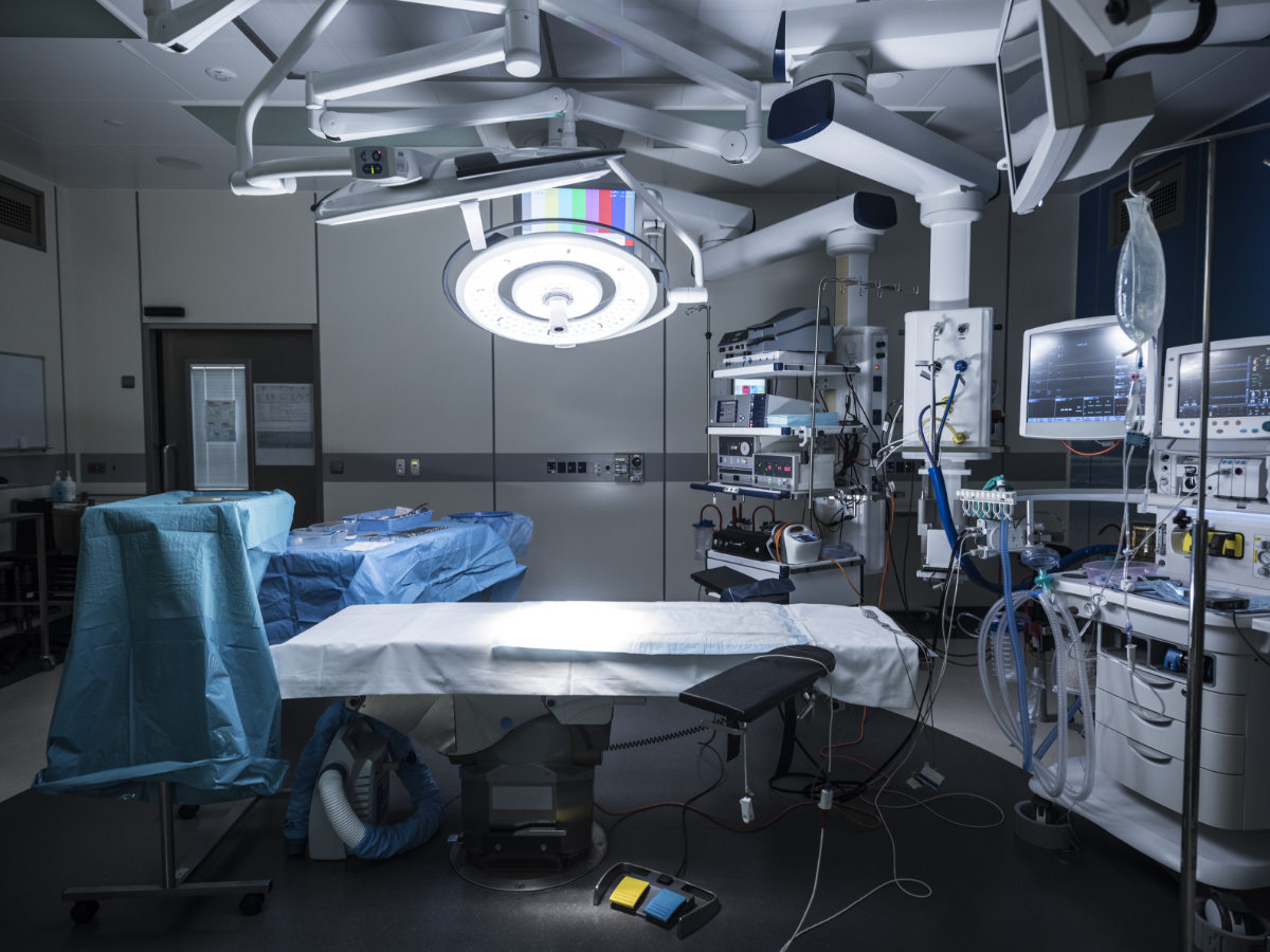 A modern operating room featuring a central surgical table covered with a white sterile sheet, surrounded by advanced medical equipment including patient monitors, anesthesia machines, and illuminated overhead surgical lights. Blue sterile drapes cover nearby instrument tables, and IV stands, tubing, and foot pedals are positioned for immediate use. The room is clean, organized, and designed for precision and safety in surgical procedures.