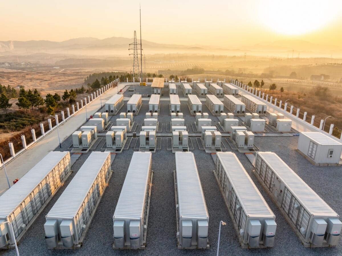 A large outdoor energy storage facility with rows of white container-like battery units at sunrise, surrounded by hills and power lines.