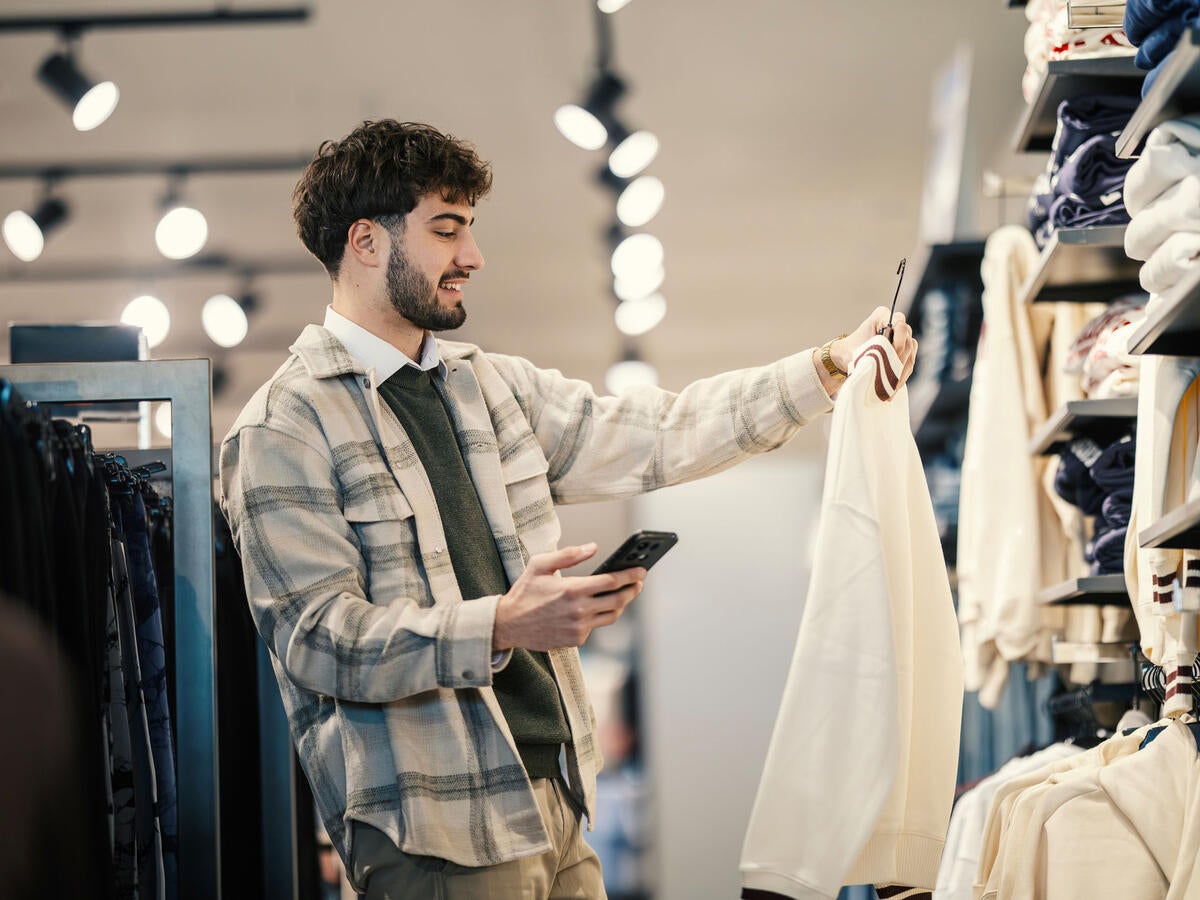  Giovane adulto che controlla il prezzo su smartphone in un negozio di abbigliamento - Foto stock Young man in a modern clothing store holds a cream sweater on a hanger, smiling as he checks his smartphone to compare prices, scan a qr code, or shop online