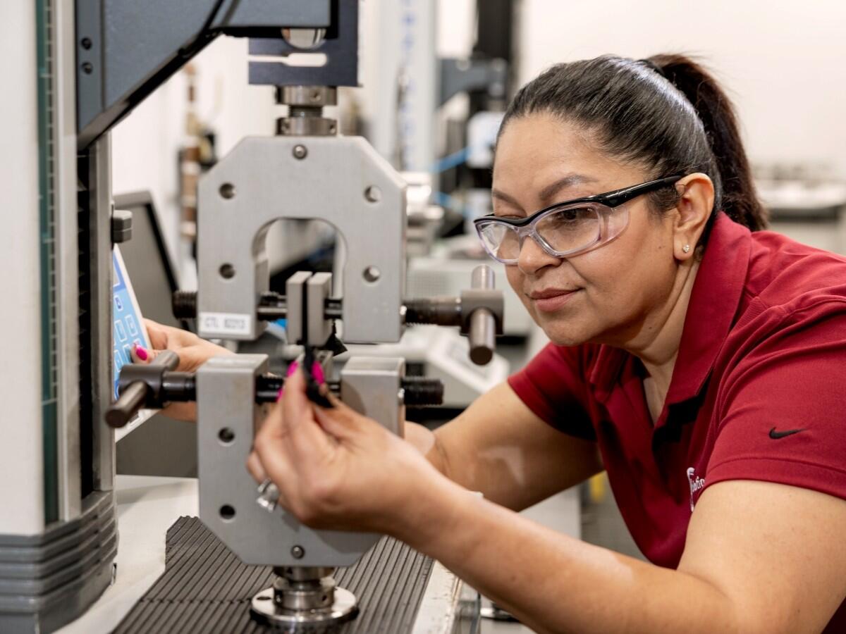 A laboratory professional wearing safety glasses adjusts a materials-testing machine.