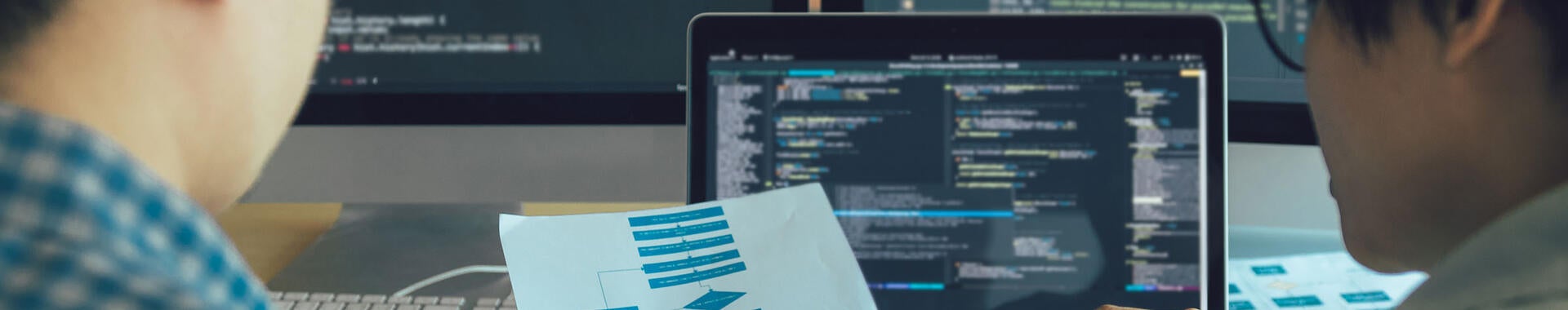 Two engineers examining a process flow on paper with coding on computer screens in the background.