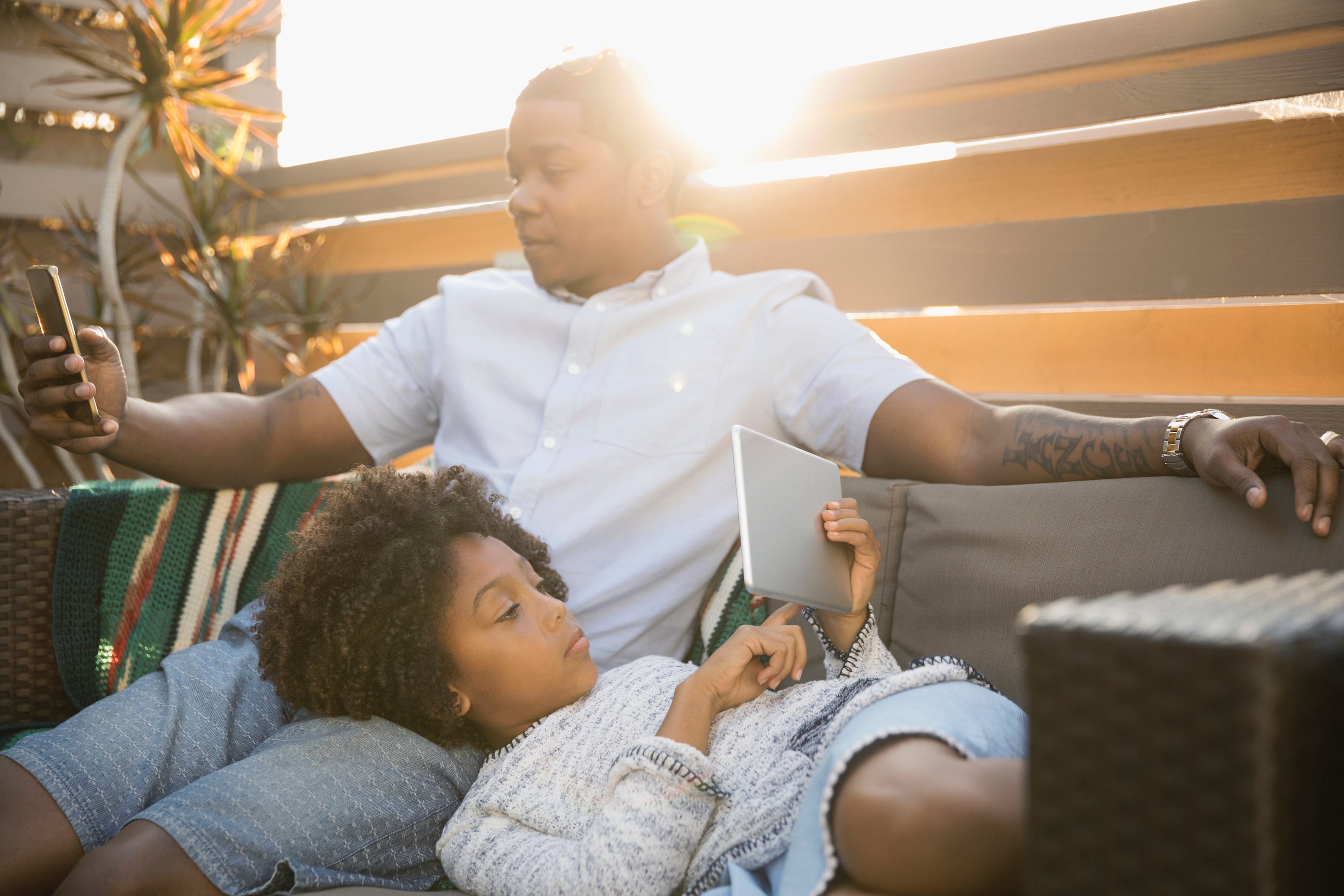 African American father and daughter using digital tablet and smart phone on sunny deck sofa