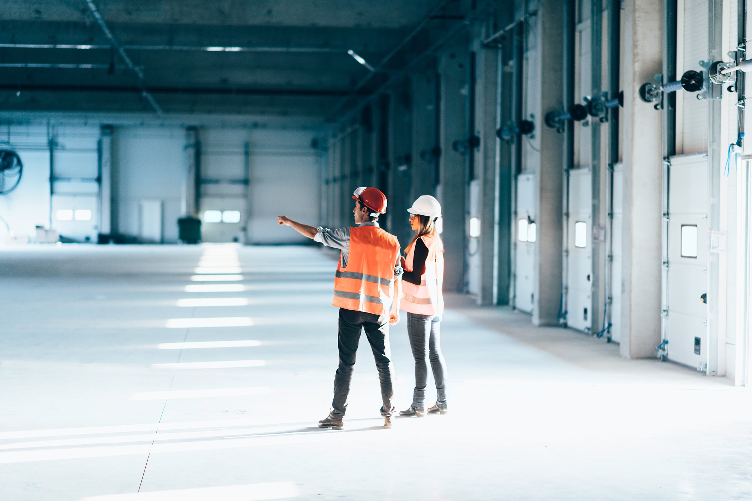 Two people in a warehouse wearing construction hats and vests pointing and looking at something