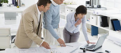 Three people in an office confer over computer and floor plans.