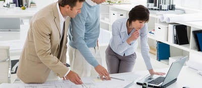Three businesspeople in an office look at a laptop and blueprints