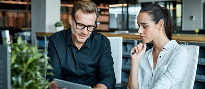 man and woman sitting next to one another at table looking at a tablet