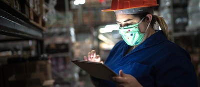 Portrait of mid adult woman wearing face mask using digital tablet - working at warehouse/industry.