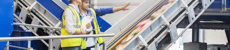 Two workers in a recycling facility stand next to moving conveyor belts.