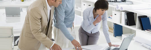 Three people in an office confer over computer and floor plans.