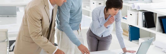 Three businesspeople in an office look at a laptop and blueprints