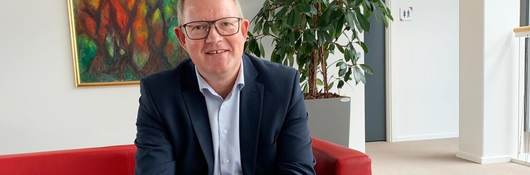 Morten Lassen casually sits on a red sofa in his Denmark office. He has on a blue suit, no tie with his hands clasped.