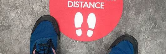 Two feet stand in front of a floor graphic that says stay safe and keep your distance