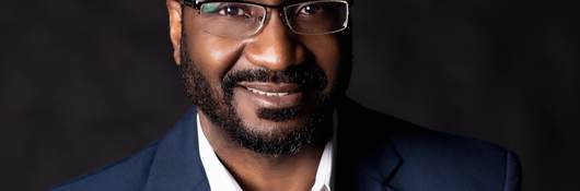Dwayne Sloan poses for headshot wearing a navy blue jacket, white collar, unbuttoned shirt on a dark background.