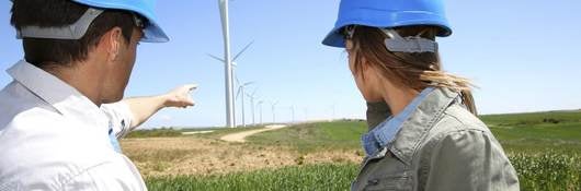 Technicians in blue hard hats standing in a field with an iPad while pointing at wind turbines