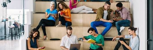 Group of young adults sitting on indoor tiered seating using their mobile phones and tablets.