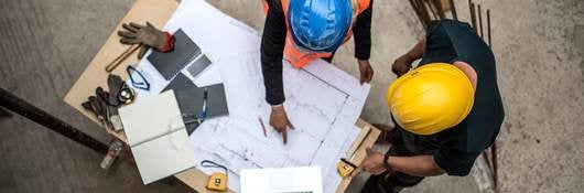 Two construction workers examine blueprints with laptop open on a project table