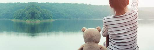A girl sitting at a lake with a stuffed teddy bear next to her