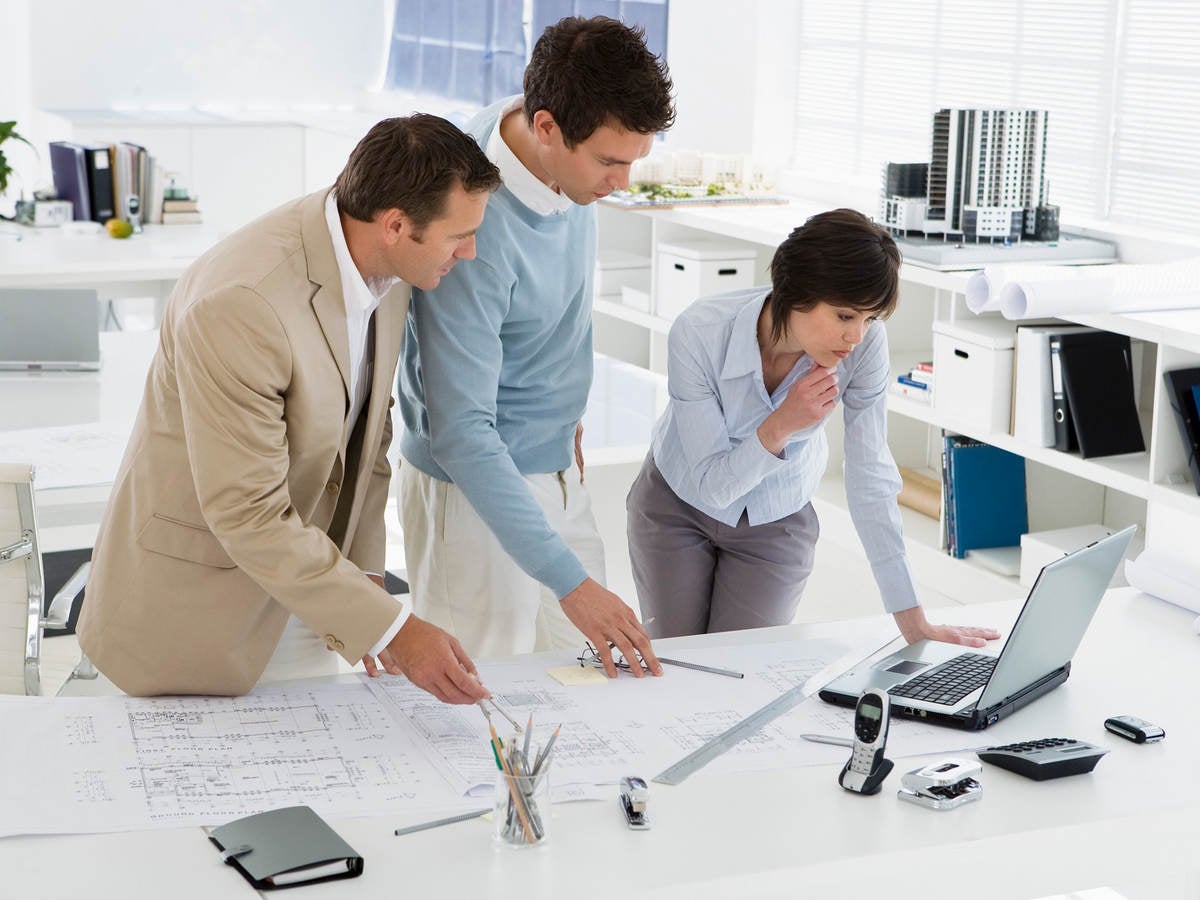 Three businesspeople in an office look at a laptop and blueprints