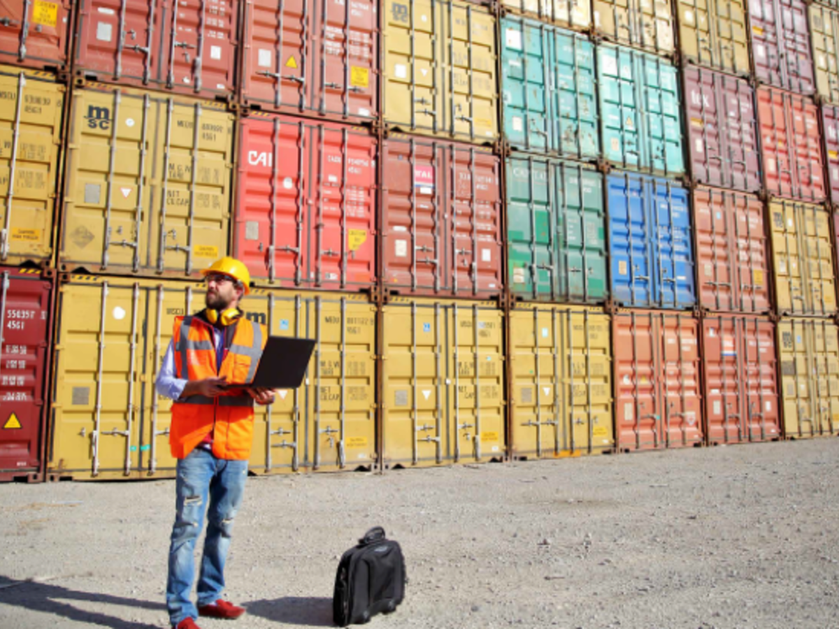 Man in orange vest/yellow hard hat in yard of shipping containers