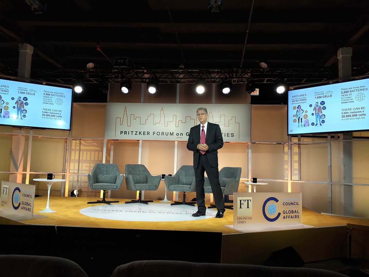 Tom Chapin, Ph.D, standing onstage with the 2019 Pritzker Forum on Global Cities banner in the center and two monitors showing the average number of batteries on any one person at any given time, to the left and right. Tom is wearing a blue suit with a red tie.