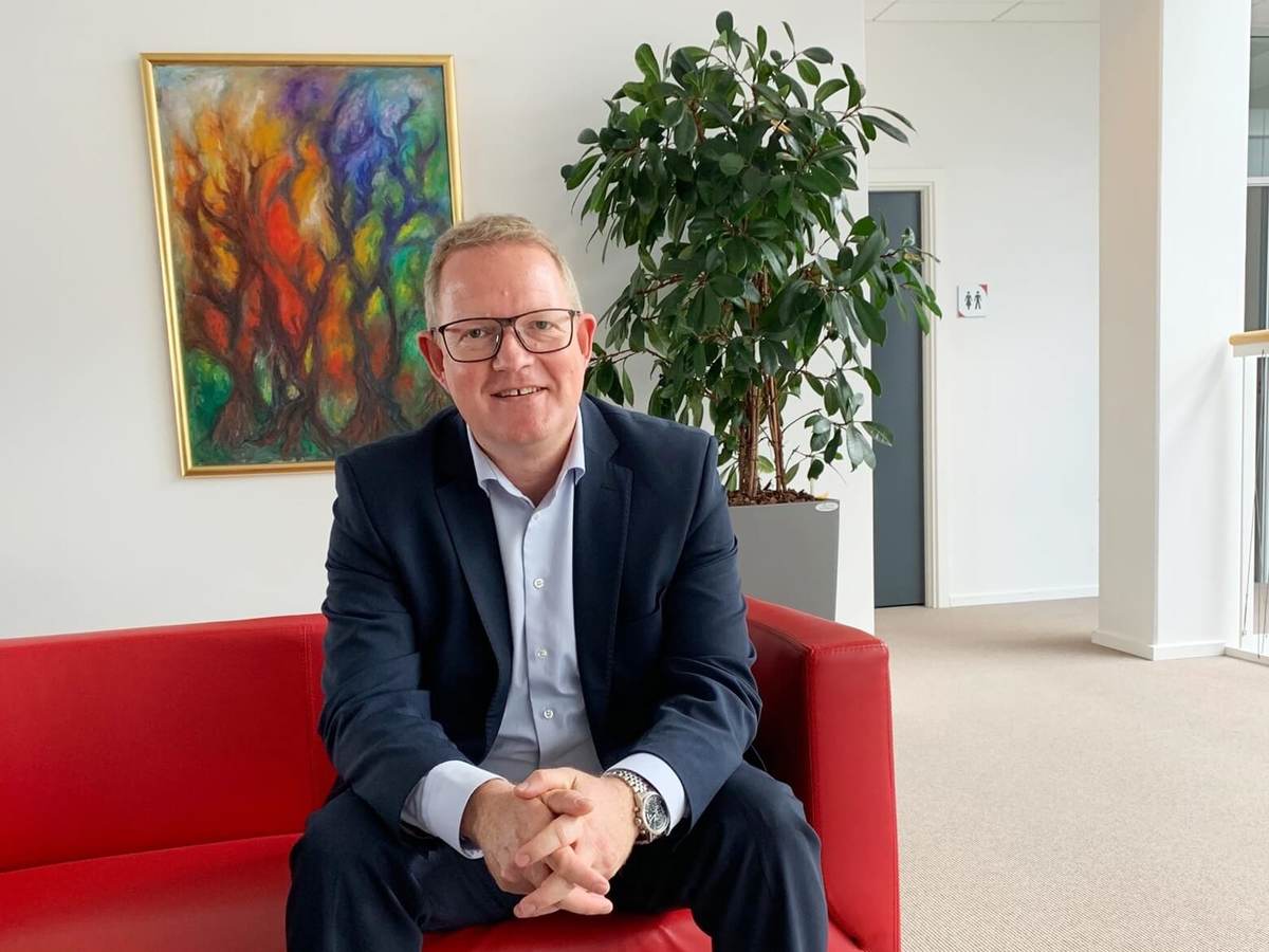 Morten Lassen casually sits on a red sofa in his Denmark office. He has on a blue suit, no tie with his hands clasped.