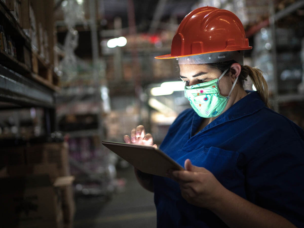 Portrait of mid adult woman wearing face mask using digital tablet - working at warehouse/industry.